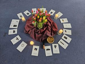 Ein Bodenarrangement aus Blumen, Kerzen und Postkarten mit den Wir-Prozess Kommunkationsempfehlungen auf blauem Teppich